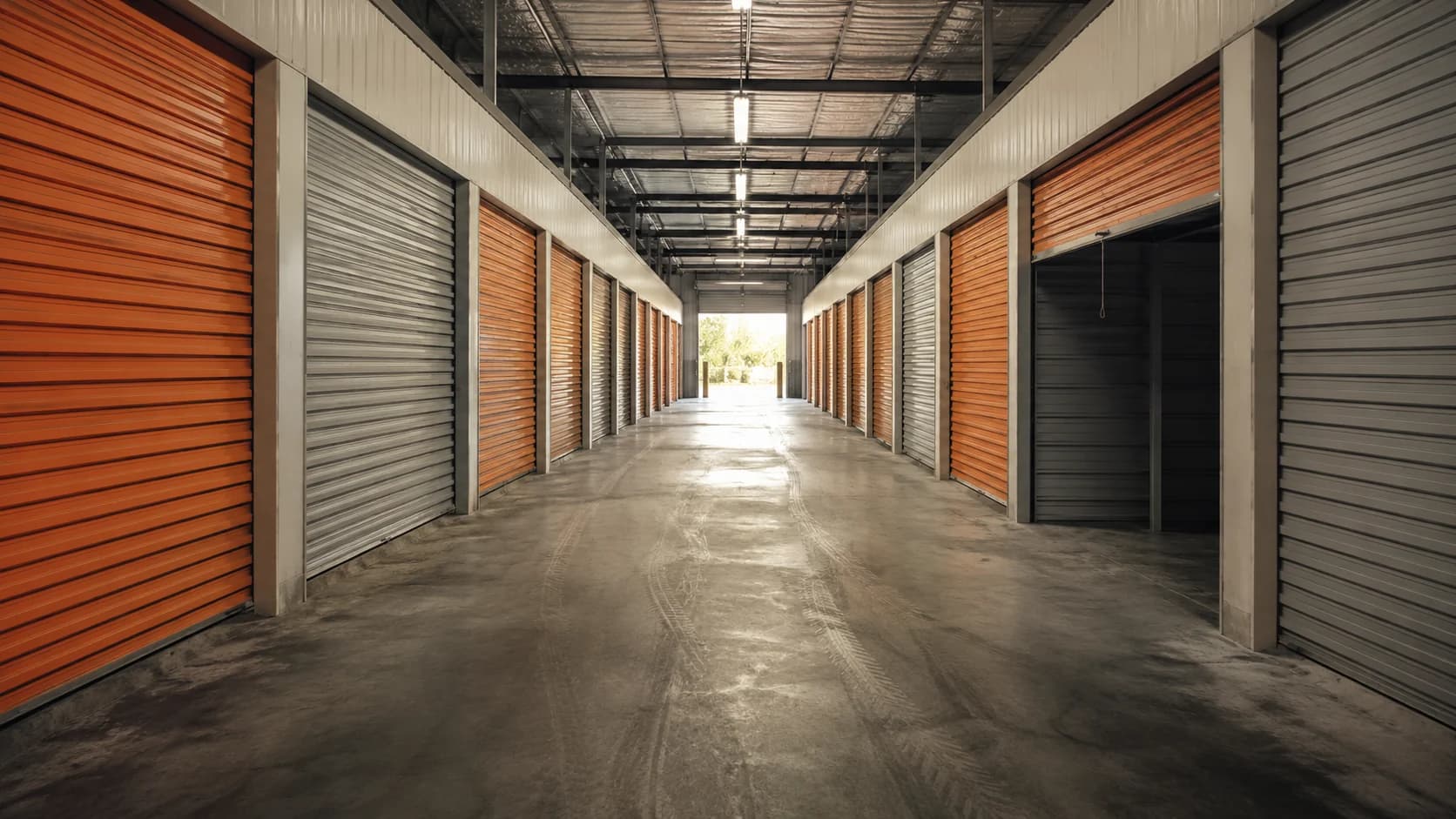 Interior corridor of a self-storage facility, orange and grey roller-shutter doors receding to a lit opening
